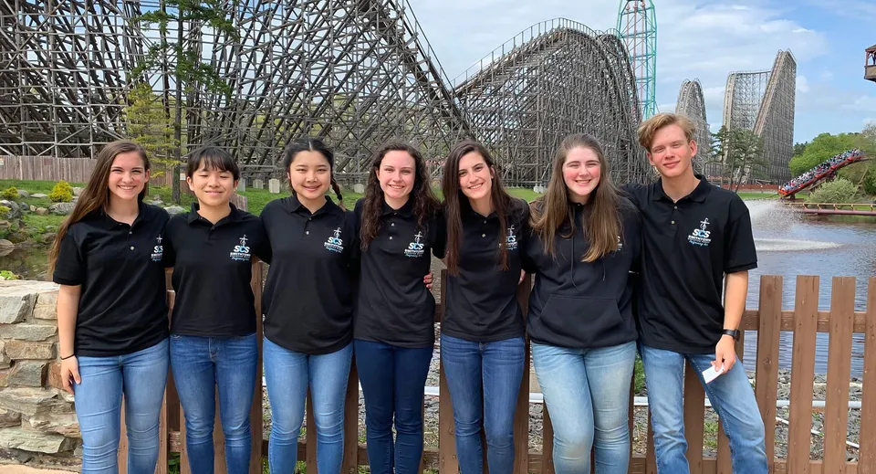 Un gruppo di sette studentesse della Smithtown Christian School è in piedi davanti a una grande montagna russa in un parco divertimenti.