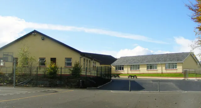 Das mehrstöckige gelb-graue Hauptgebäude von Coláiste Mhuire Ballygar vor einem bewaldeten Hinterhof und blauem Himmel.