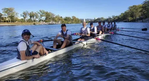 Un grupo de estudiantes de la Brisbane Grammar School remando en un río, rodeados de árboles bajo un cielo azul.