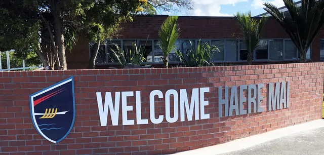 Un muro di mattoni con un grande cartello WELCOME HAEREWA nel campus dell'Howick College, circondato da alberi e aree verdi.