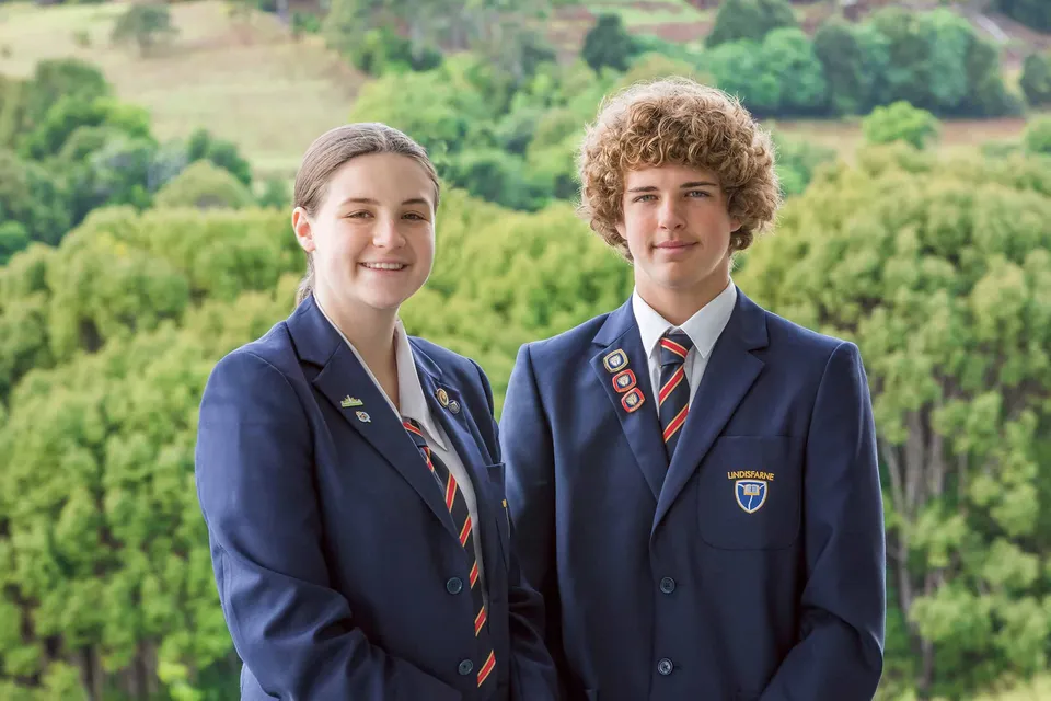 Due studenti con la divisa della Lindisfarne Anglican Grammar School posano davanti a un paesaggio verde.