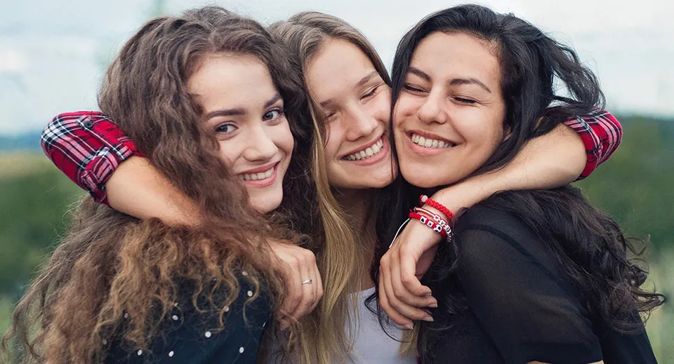 Tre studentesse della Sardis Secondary School si abbracciano ridendo all'aperto.