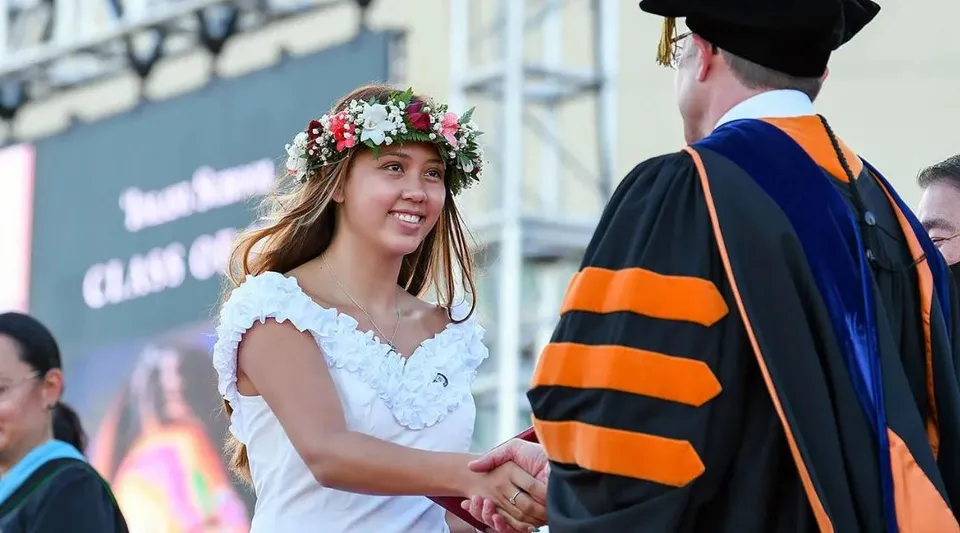 Una studentessa della Iolani School indossa un vestito bianco e una ghirlanda di fiori e stringe la mano a un uomo in abito accademico davanti a un edificio universitario.