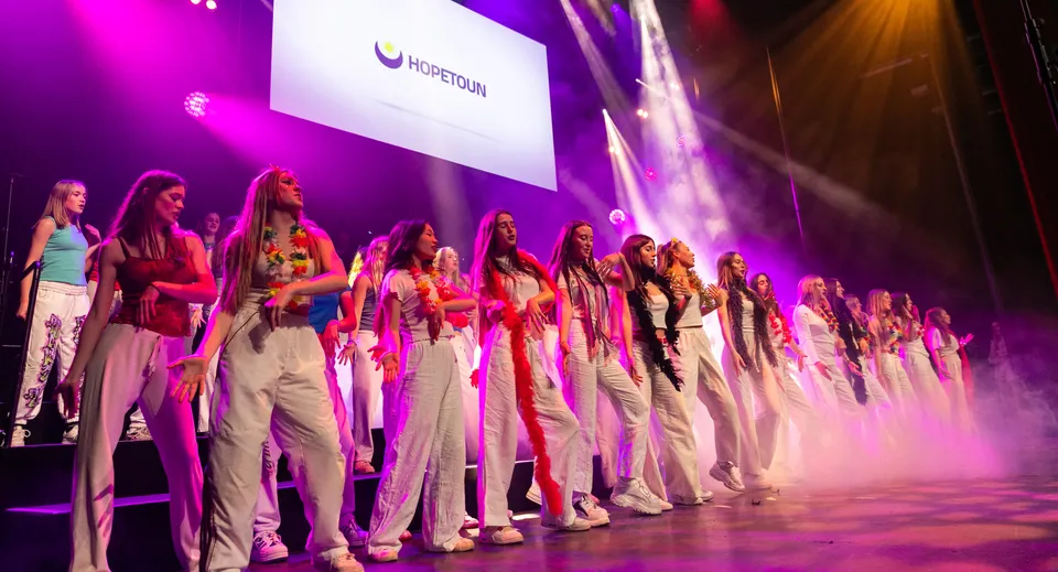 Un gruppo di donne in abiti colorati si trova sul palco della Westminster School davanti a un grande logo e a un'illuminazione colorata.