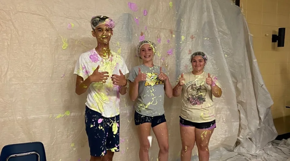 Tre studenti della Howard S. Billings High School coperti di pittura colorata si trovano in una stanza davanti a una tenda bianca.