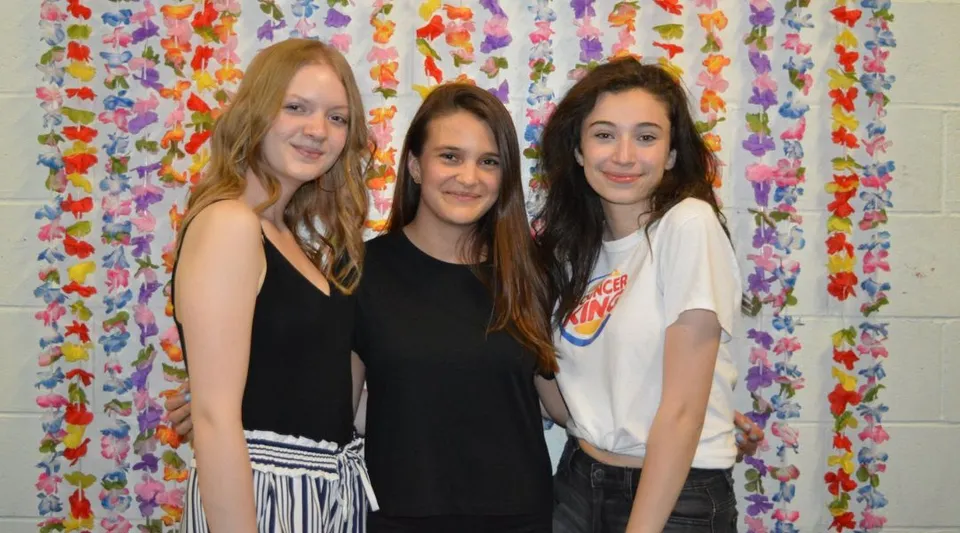 Tre studentesse della St. Thomas High School sono in piedi davanti a una tenda di fiori variopinta.