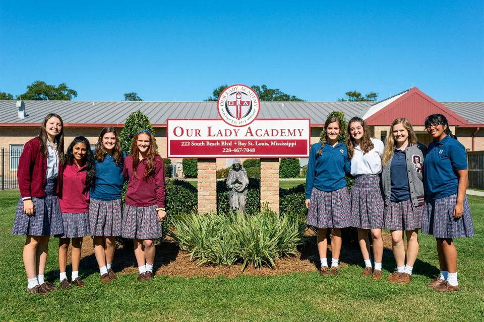Otto studentesse della Our Lady Academy in divisa scolastica posano davanti al cartello della scuola.