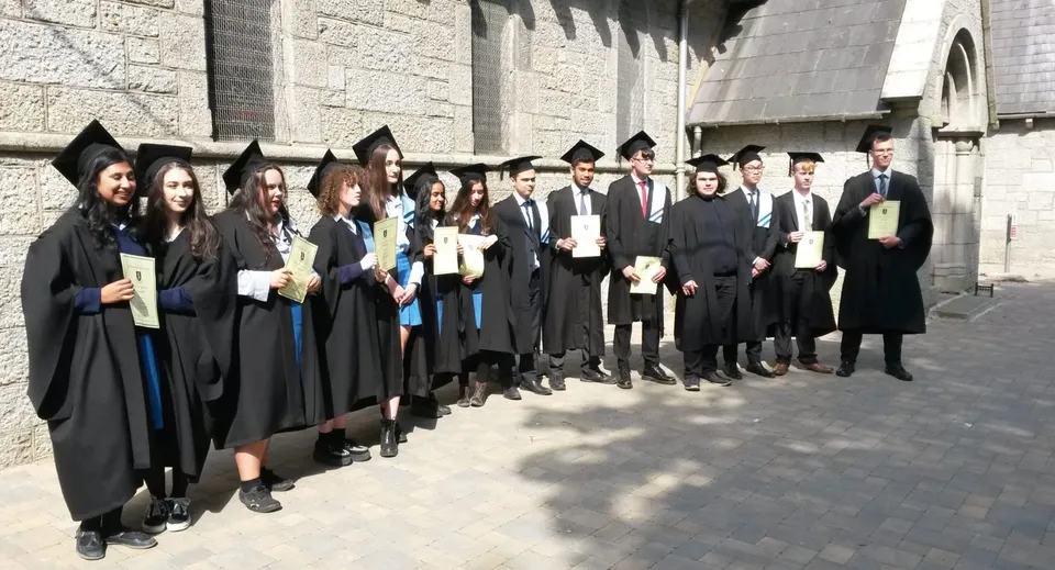 Un gruppo di diplomati e diplomate della John Scottus Secondary School è in piedi in abito accademico davanti a un edificio in pietra.