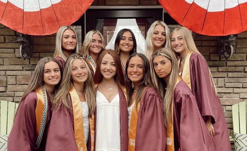 Un gruppo di diplomate della Central Regional High School indossa toghe bordeaux e sta davanti a un edificio in mattoni con decorazioni rosse e bianche.