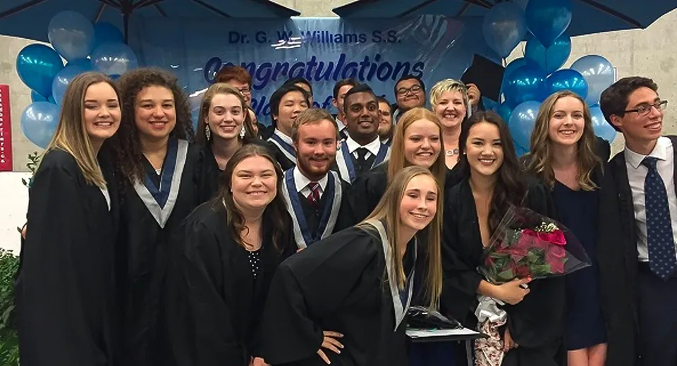 Studenti della Dr. G.W. Williams Secondary School si radunano in toga da diploma davanti a uno striscione di auguri con un ombrello blu sopra.