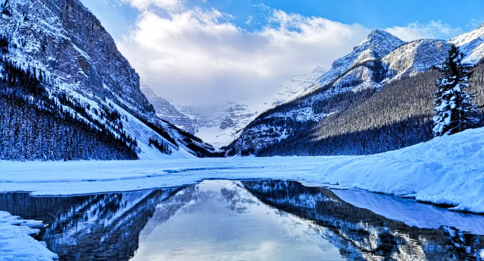 Montagne innevate si riflettono in un lago parzialmente ghiacciato sotto un cielo azzurro.