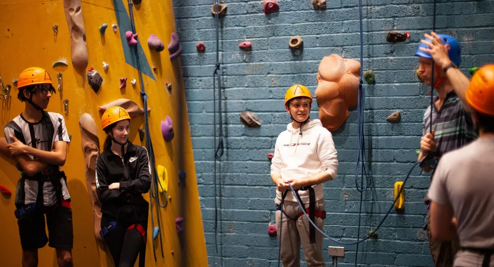 Studenti della Westminster School si arrampicano con imbracature ed elmetti su una parete d'arrampicata colorata in una palestra d'arrampicata.