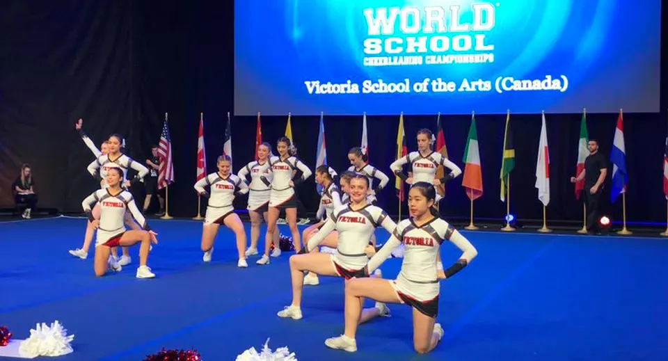 Un gruppo di cheerleader in uniforme bianca si esibisce sul palco della Victoria School of the Arts davanti alle bandiere di diversi paesi.