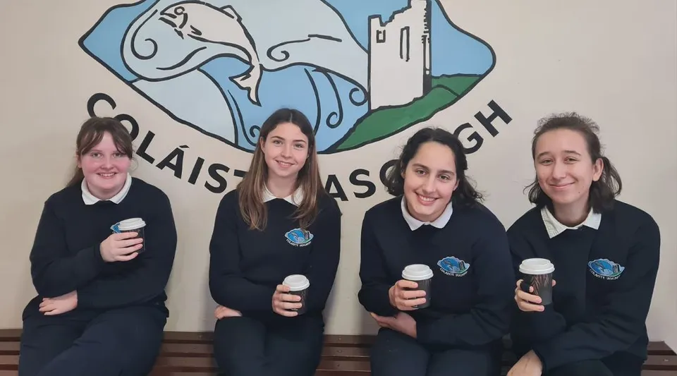 Quattro studenti in divisa scolastica sono in piedi davanti a un murale con un castello e un paesaggio nel campus dell'Iascaigh College.