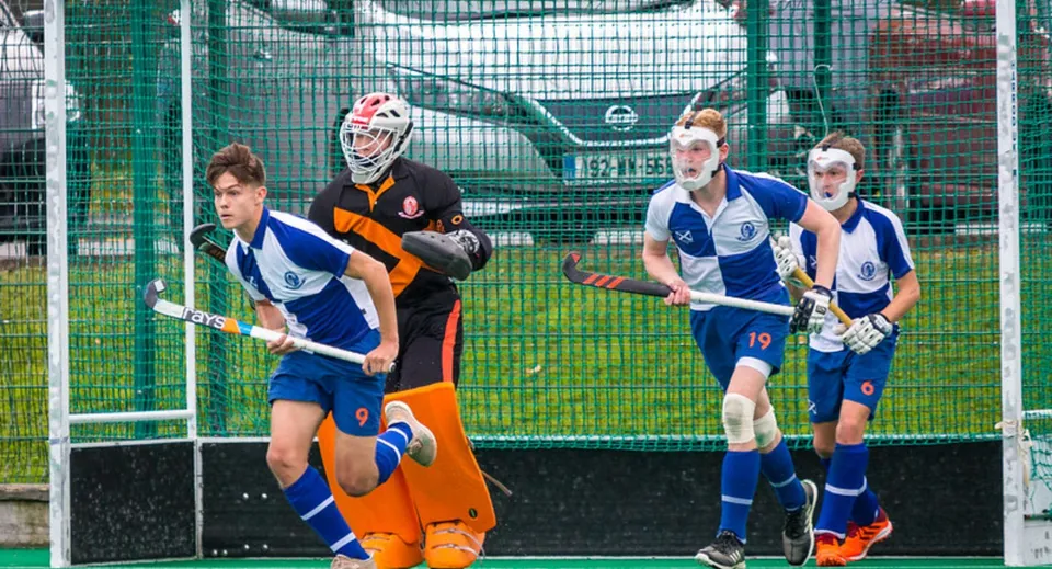 Un gruppo di giovani giocatori di hockey in maglia con un portiere davanti a una rete metallica verde nel campus del St. Andrew's College.