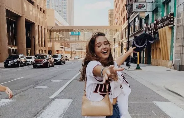 Chica sonriente vestida con camiseta blanca extendiendo el brazo en una calle de la ciudad