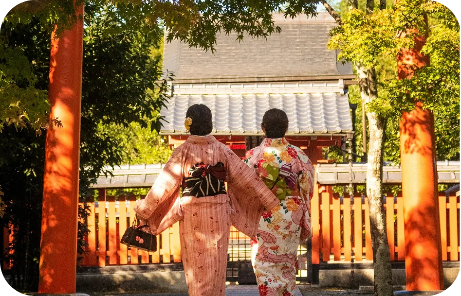Zwei junge Frauen in Kimonos gehen von hinten gesehen auf ein Tempeltor mit roten Säulen zu