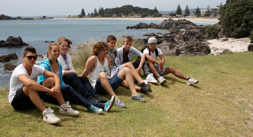 Un gruppo di studenti della Whakatane High School seduti sull'erba vicino alla costa rocciosa, con acqua ed edifici sullo sfondo.