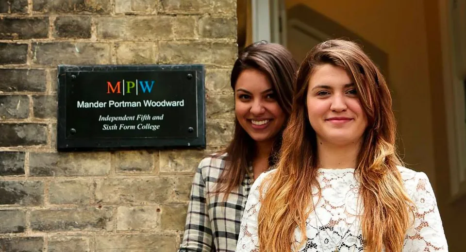 Due studentesse con i capelli lunghi e scuri stanno davanti a una parete in mattoni con l'insegna del MPW Cambridge.