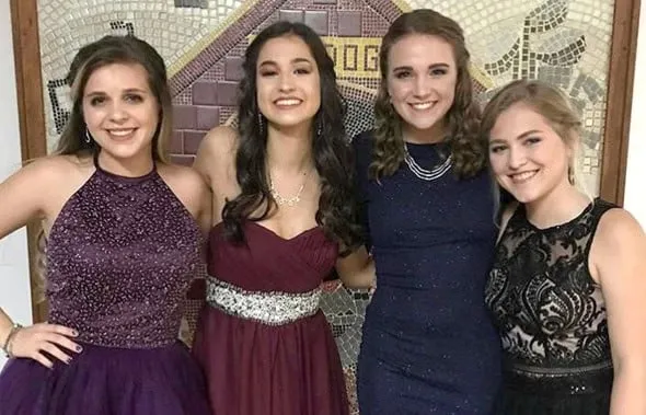 Cuatro chicas sonrientes con vestidos de fiesta elegantes posan juntas frente a un mosaico
