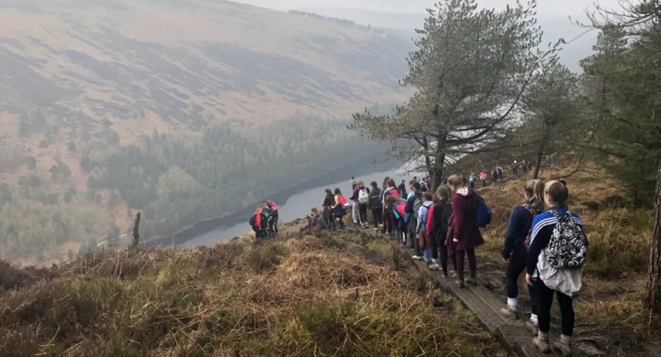 Un gruppo di studenti e studentesse della Cornafulla National School fa un'escursione su un sentiero attraverso un paesaggio boschivo di montagna con un fiume sullo sfondo.