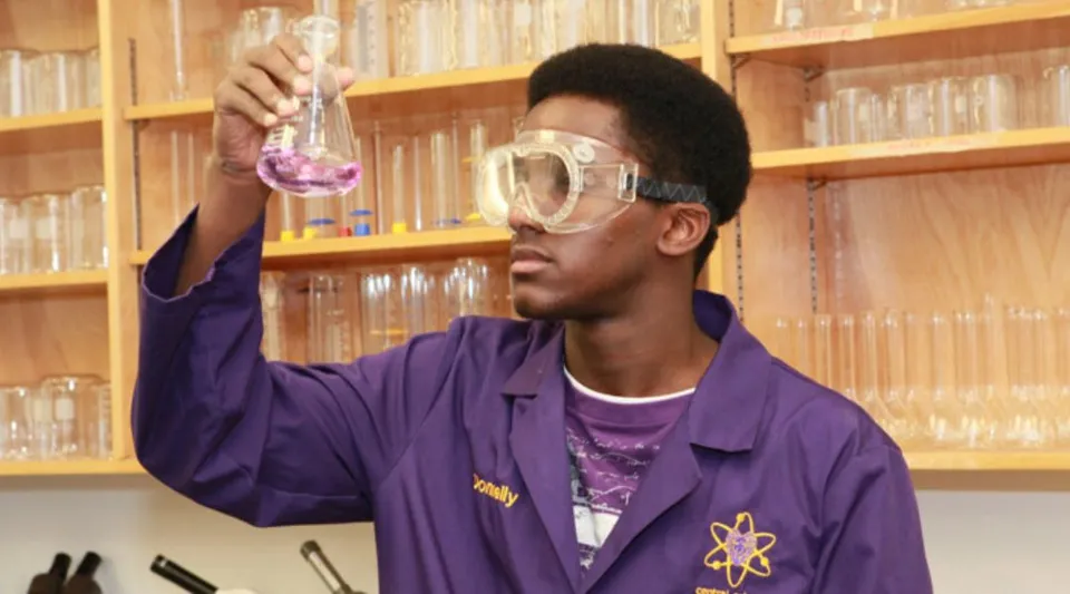 Uno studente della Crescent Heights High School tiene in alto in un laboratorio un beaker con un liquido rosa davanti a una libreria.