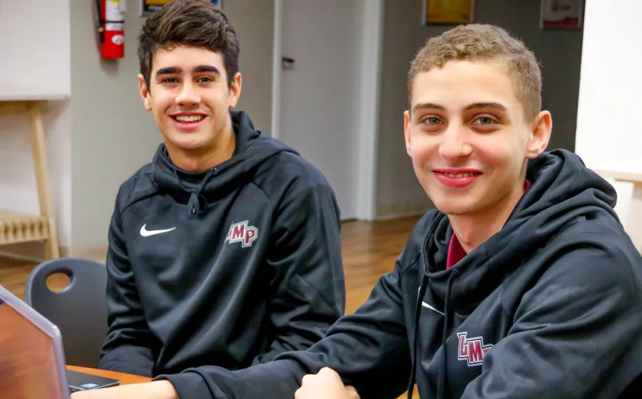 Dos alumnos de la Lake Mary Preparatory School llevan sudaderas con capucha oscuras con el logo de UH y sonríen en un interior.