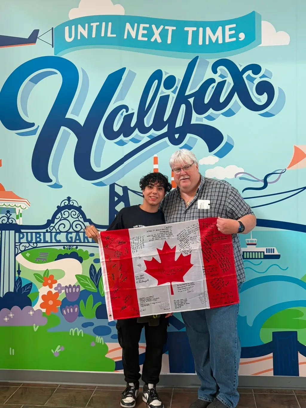 Un ragazzo e un uomo mostrano una bandiera canadese firmata davanti al murale 'Until next time, Halifax'.