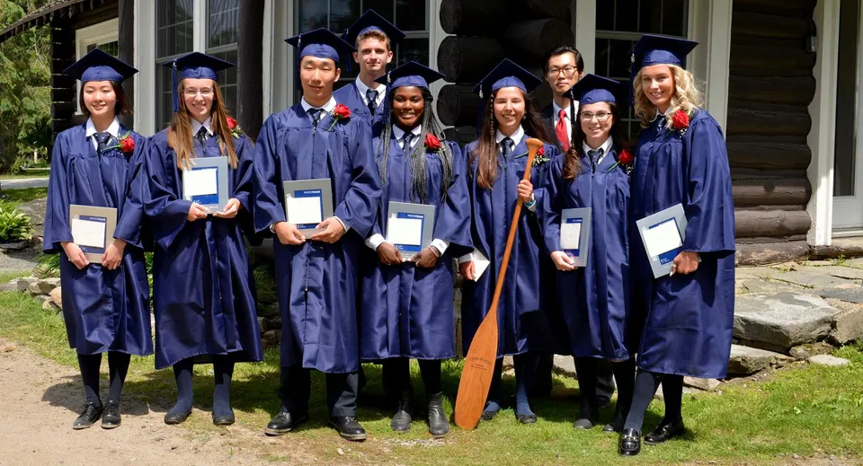 Un gruppo di diplomati con toghe blu mostra i propri diplomi davanti a un edificio in legno nel campus del Rosseau Lake College.