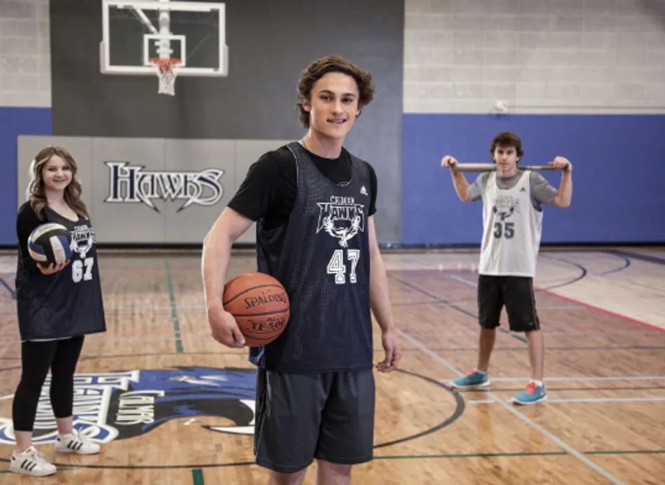 Tre studenti della Calmar Secondary School con un pallone da basket, uno da pallavolo e una mazza da baseball in palestra.