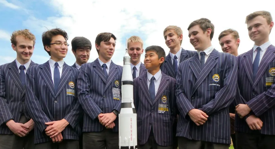 Un gruppo di giovani uomini del Sheldon College posa in abito blu scuro davanti a un cielo nuvoloso.