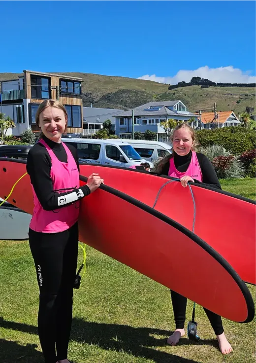 Joseline und eine Freundin im Neoprenanzug halten ein rotes Surfbrett in Neuseeland