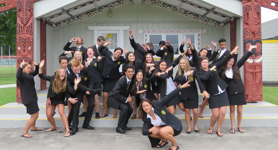 Un gruppo di diplomati della Whakatane High School festeggia davanti a una tradizionale casa di riunione Maori con intagli.