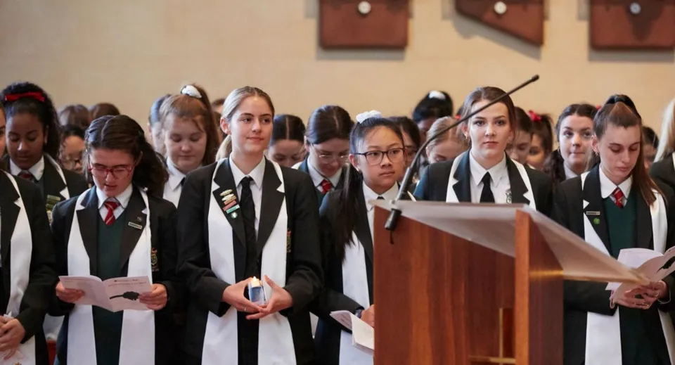 Studenti del Mercedes College in abiti formali si trovano in una stanza con pannelli di legno vicino al podio.