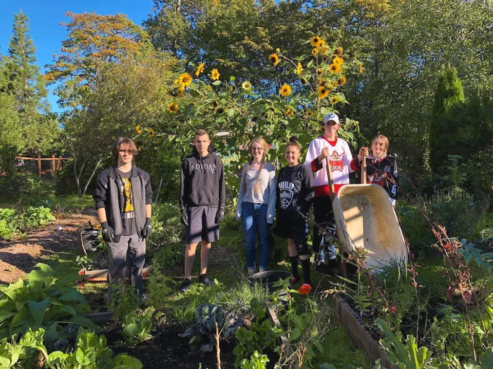 Un gruppo di persone si trova in un giardino colorato con alberi e una scultura in pietra nei pressi della John Barsby Secondary School.