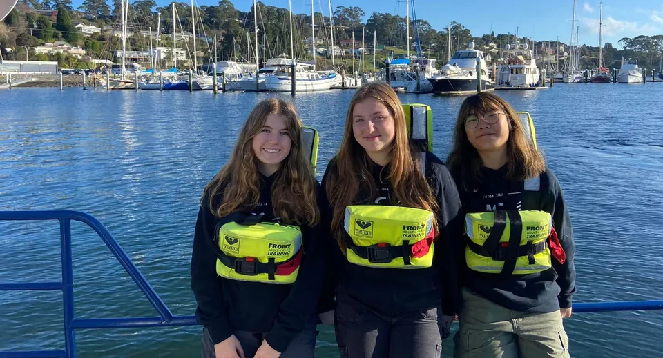 Tre studentesse della Le Fevre High School indossano giubbotti di salvataggio gialli davanti a un porto con barche.