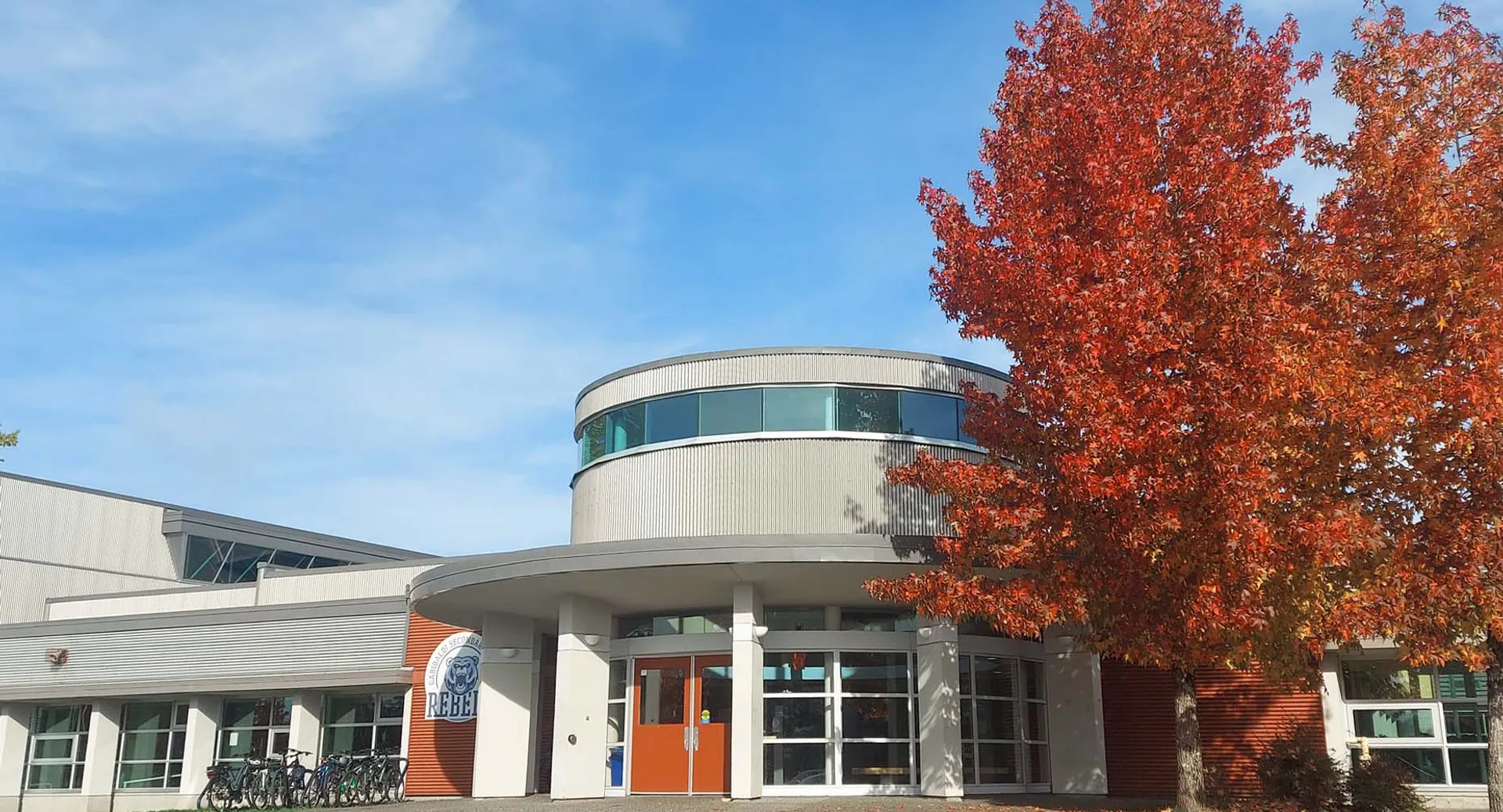 L'edificio moderno della Garibaldi Secondary School con pareti in vetro si trova davanti ad alberi autunnali rossi sotto un cielo blu.