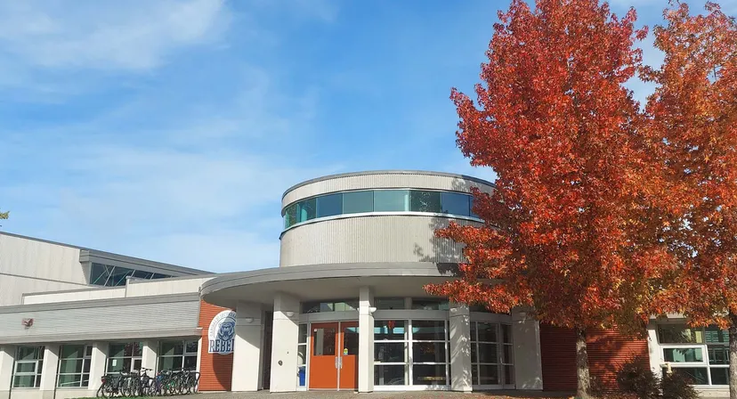 L'edificio moderno della Garibaldi Secondary School con pareti in vetro si trova davanti ad alberi autunnali rossi sotto un cielo blu.