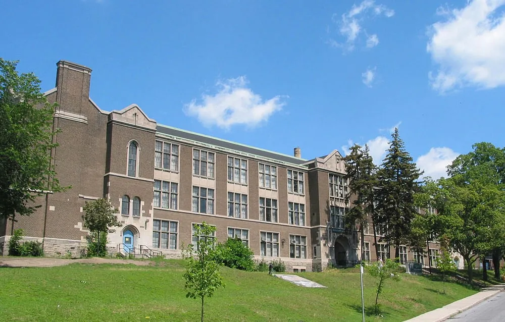 Un edificio de ladrillo de varios pisos de la Western Technical-Commercial School se encuentra rodeado de árboles y un primer plano cubierto de hierba bajo un cielo azul.