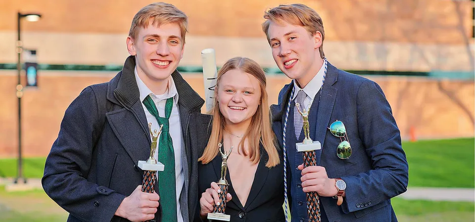 Tre studenti della Grand Rapids Christian High School sono insieme all'aperto davanti a una staccionata di legno e ad alcuni alberi.