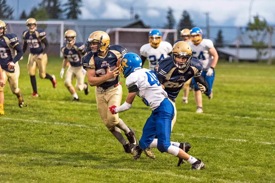 Un gruppo di giocatori di football della Salmon Arm Secondary gioca con intensità su un campo d'erba con spettatori sullo sfondo.