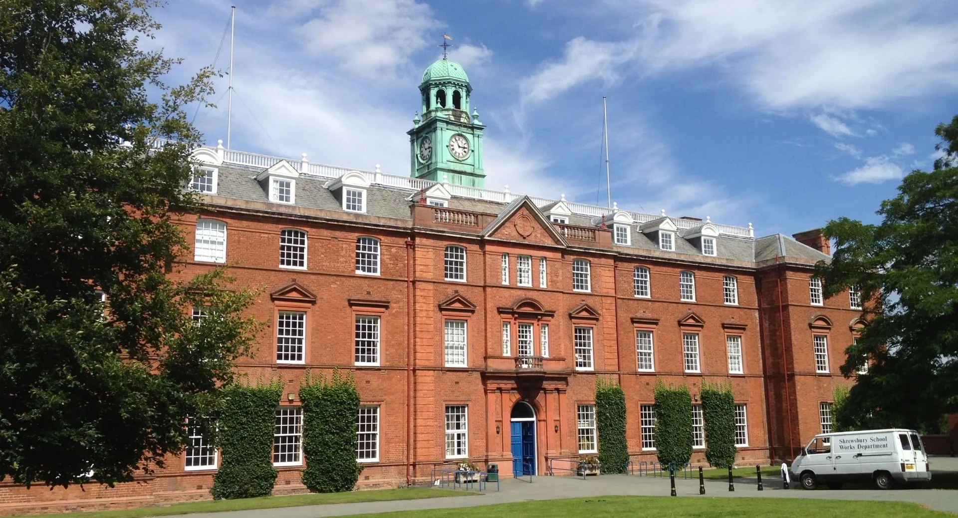 L'edificio storico in mattoni con il tetto della torre verde si trova nel campus della Shrewsbury School sotto un cielo blu.