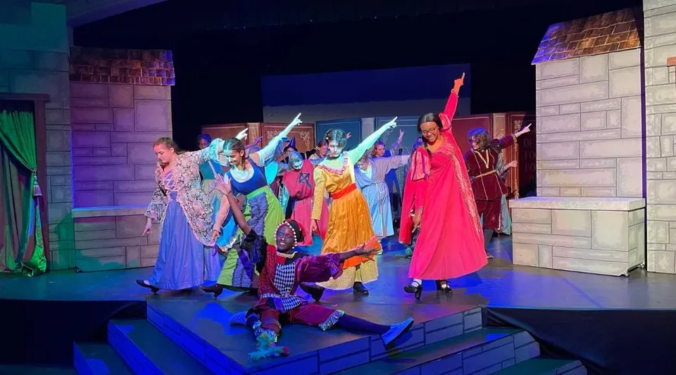 Un gruppo di attori in abiti colorati si trova sul palco della Laurel School davanti a una scenografia architettonica con un'illuminazione vivace.