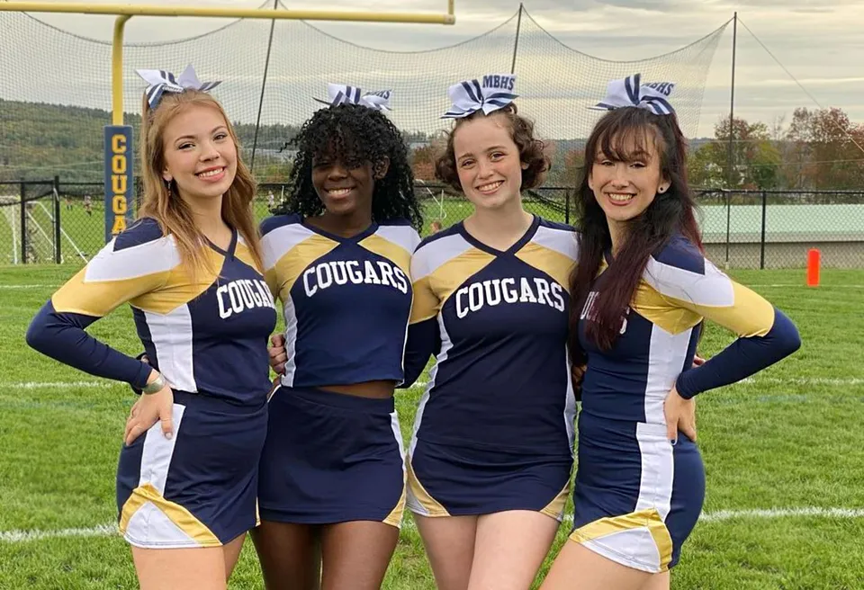 Quattro studentesse della Mount Blue High School posano insieme sul campo sportivo con le montagne sullo sfondo.