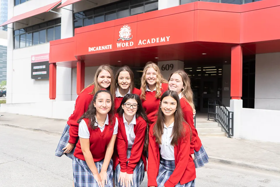 Un gruppo di giovani donne è in piedi in toghe rosse da diploma davanti all'edificio della Incarnate Word Academy.