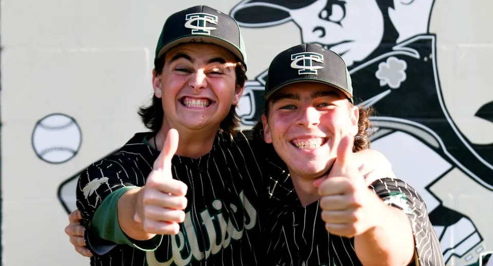 Due studenti della Trinity Catholic High School indossano cappellini da baseball e maglie sportive e mostrano il pollice in su davanti a un murales con un personaggio dei fumetti.