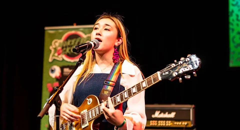Una studentessa dell'ACG Sunderland canta e suona la chitarra durante un'esibizione sul palco davanti a uno sfondo colorato.