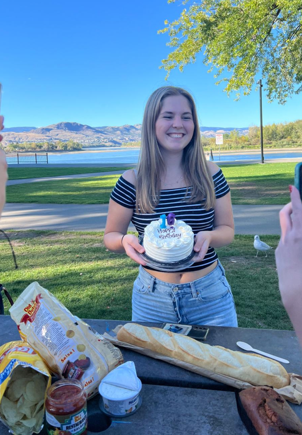 Studentessa sorridente tiene una torta di compleanno a un picnic all'aperto vicino a un lago