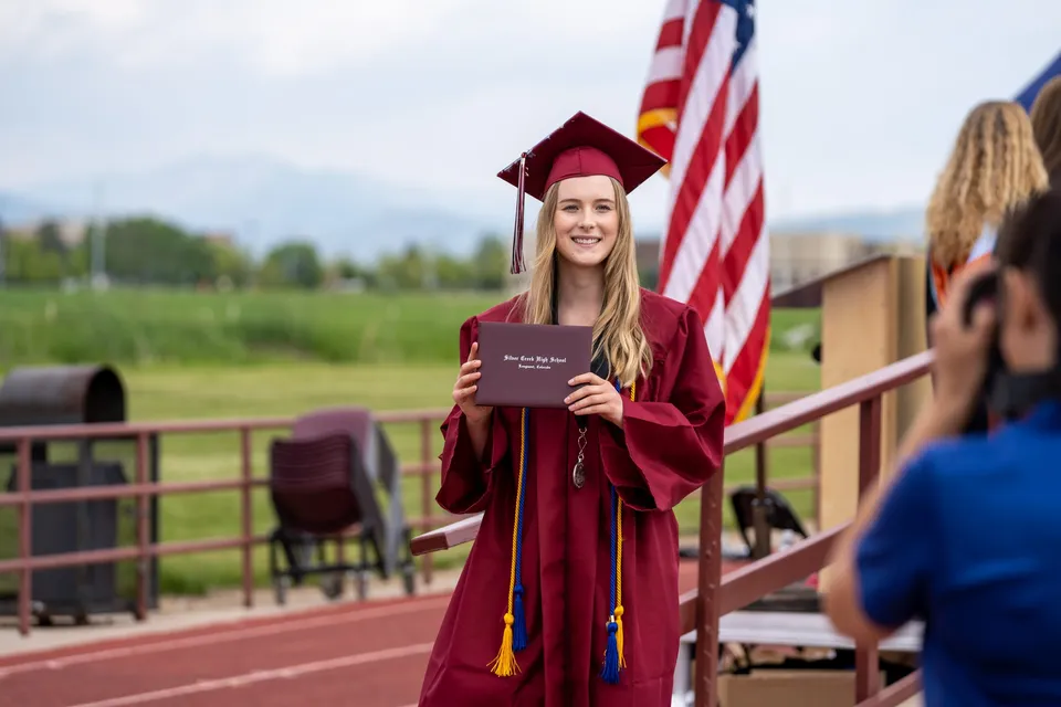 Una giovane donna in toga rossa da diploma si trova sul balcone della Red Creek High School con vista su montagne e alberi.