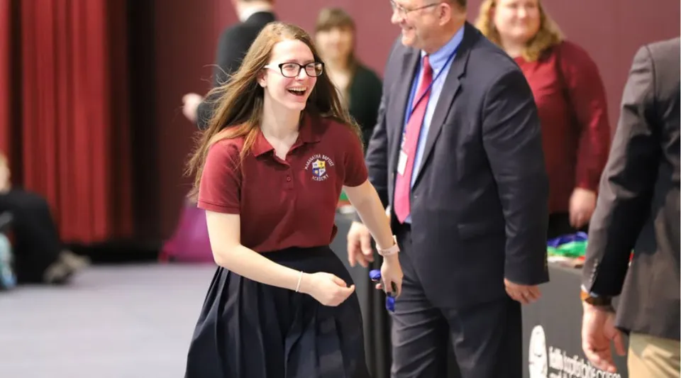 Una studentessa della Maranatha Baptist Academy sorride con gli occhiali davanti a degli adulti in abito formale.